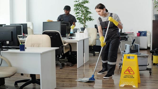 Young black man wiping computer monitors while Caucasian woman in coveralls and yellow gloves with mop cleaning floor in office